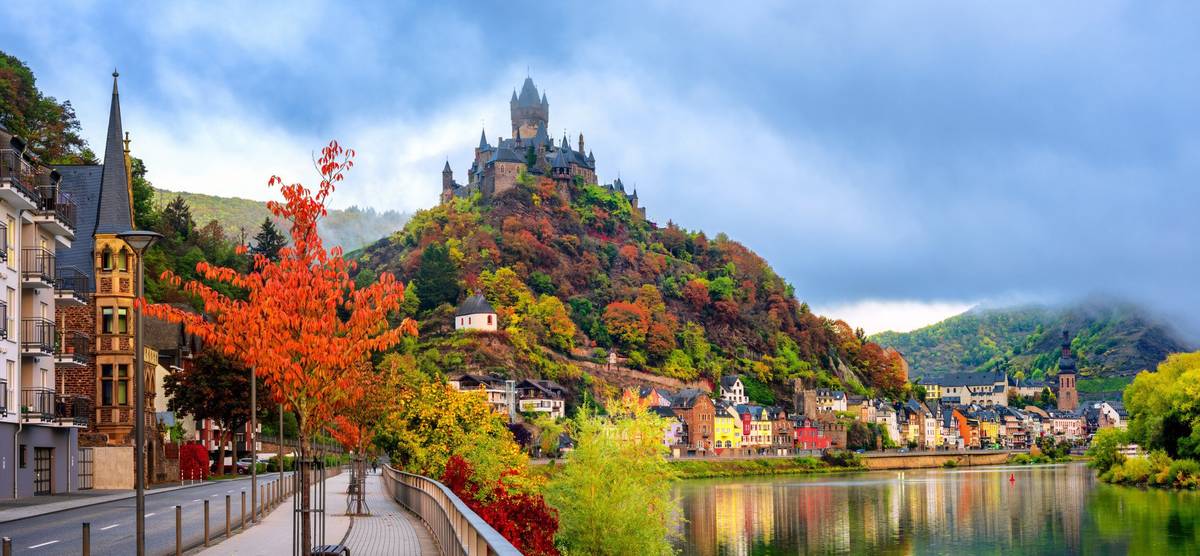 Blick auf eine Stadt am Fluss mit historischen Gebäuden und einer Burg auf einem hohen Hügel inmitten herbstlich gefärbter Landschaft