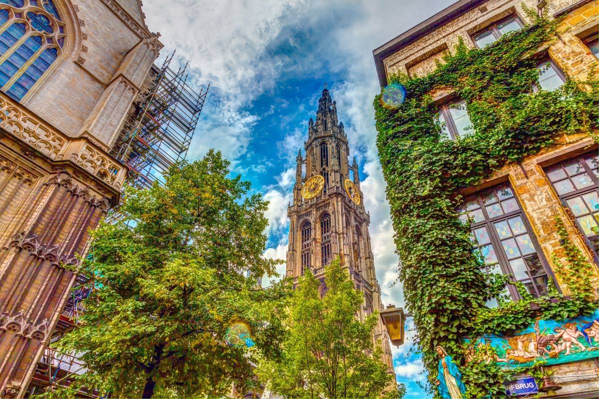 Blick von unten nach oben auf Turmuhr mit goldenen Ziffern und bewachsenen historischen Gebäuden bei Tag