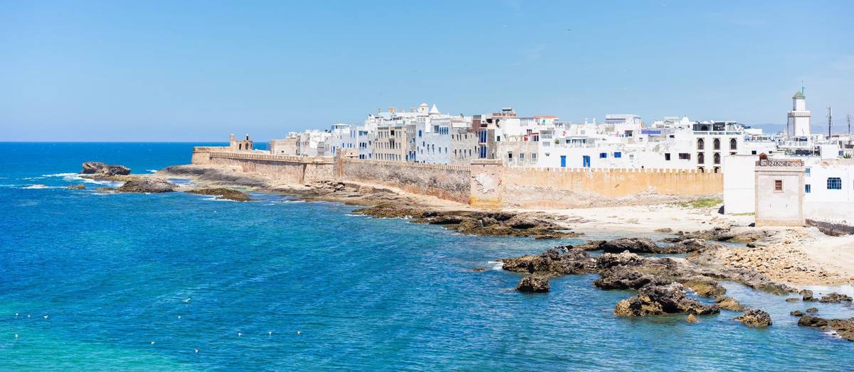 Panoramaaufnahme am Tag der Küste im marokkanischen Essaouira. Das Wasser ist tiefblau, die abgebildeten Häuserfronten weiß.