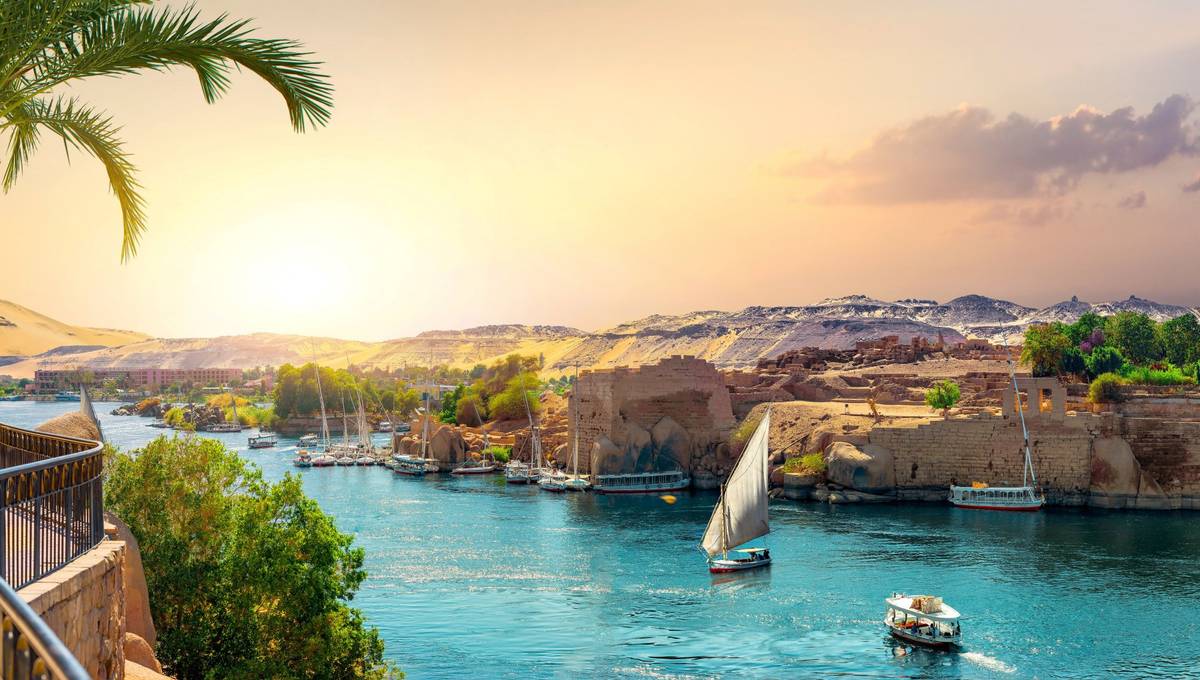 Der Fluss Nil in Ägypten als Panoramaaufnahme. Das Wasser ist tiefblau und eine Felukke fährt zentral auf dem Wasser, andere Boote liegen an Anlegern an.