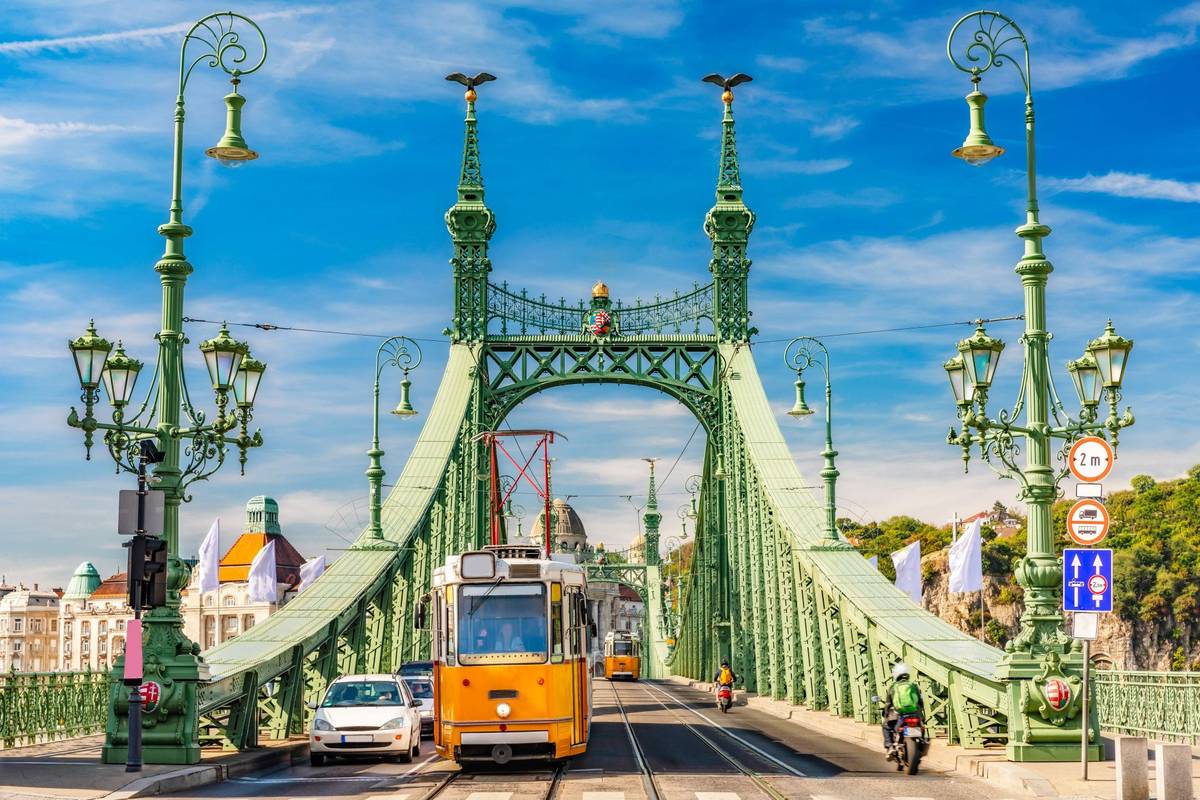Freiheitsbrücke Szabadság híd in Budapest bei Tag mit grüner Eisenkonstruktion, Jugendstil-Ornamenten und goldenen Turul-Vögeln auf den Pylonen. Gelbe Bahnen fahren auf den Schienen