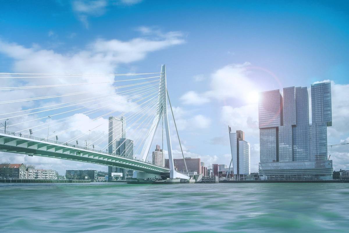 Strahlend helle Aufnahme von der Erasmusbrücke in Rotterdam. Das Wasser des Flusses ist türkisfarben und die Skyline in einem hellen metallischen Grau zu sehen.