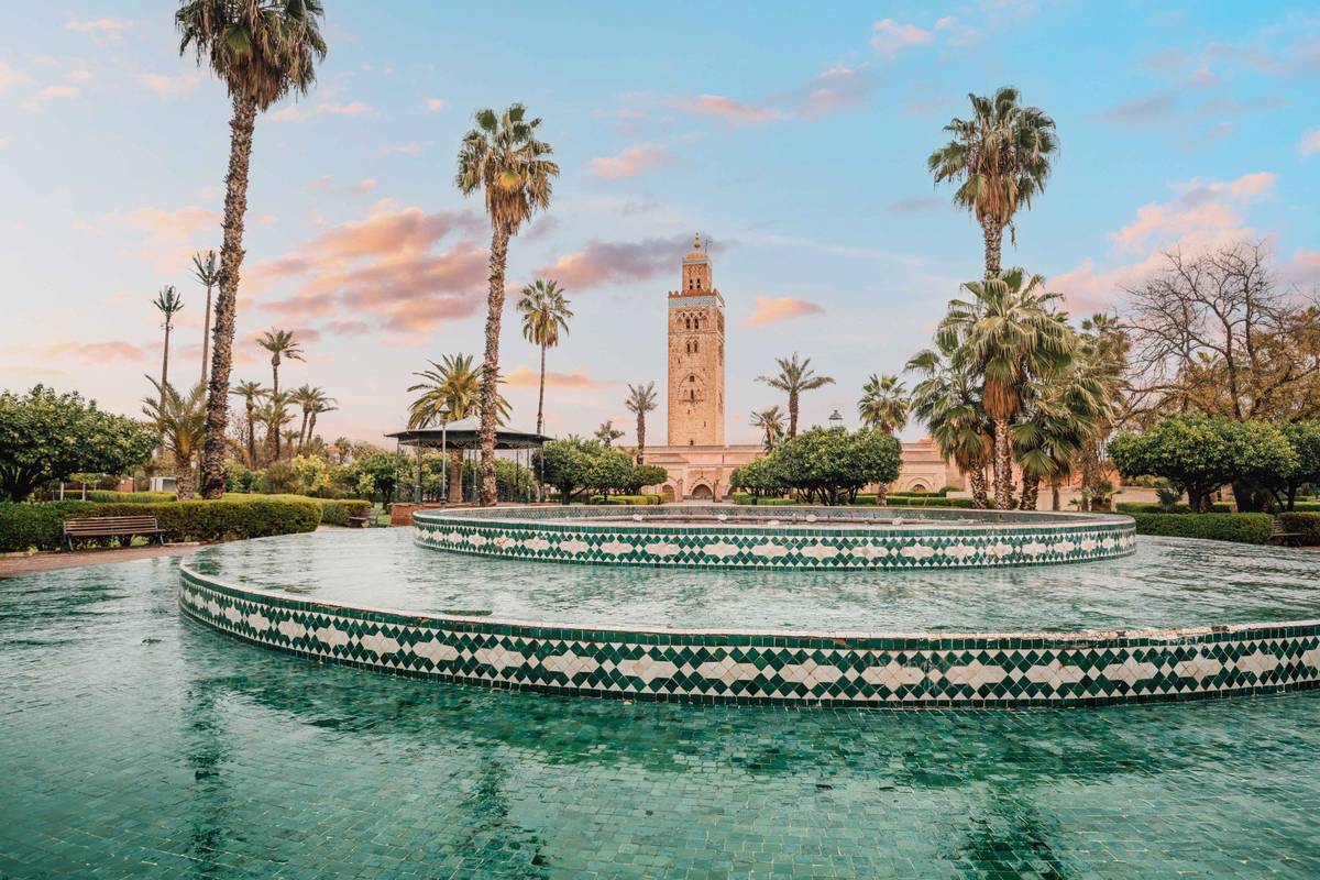 Aufnahme eines Brunnens mit grünem Mosaik vor Palmen im marokkanischen Marrakesch. Im Hintergrund ist die majestätische Koutobia Moschee zu sehen.