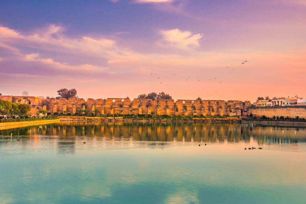 Blick auf das Becken der Norias, einem künstlichen Wasserreservoir (Sahrij Swani) in Meknes. Der Himmel ist in lila und blau getaucht mit einzelnen langgezogenen Wolken,