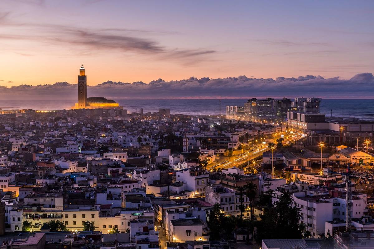 Abendliche Aufnahme von Casablanca in Marokko. Straßenlichter erhellen die Häuser. Die Hassan II. Moschee ragt in der Ferne auf, der Himmel ist grau-lila eingefärbt.