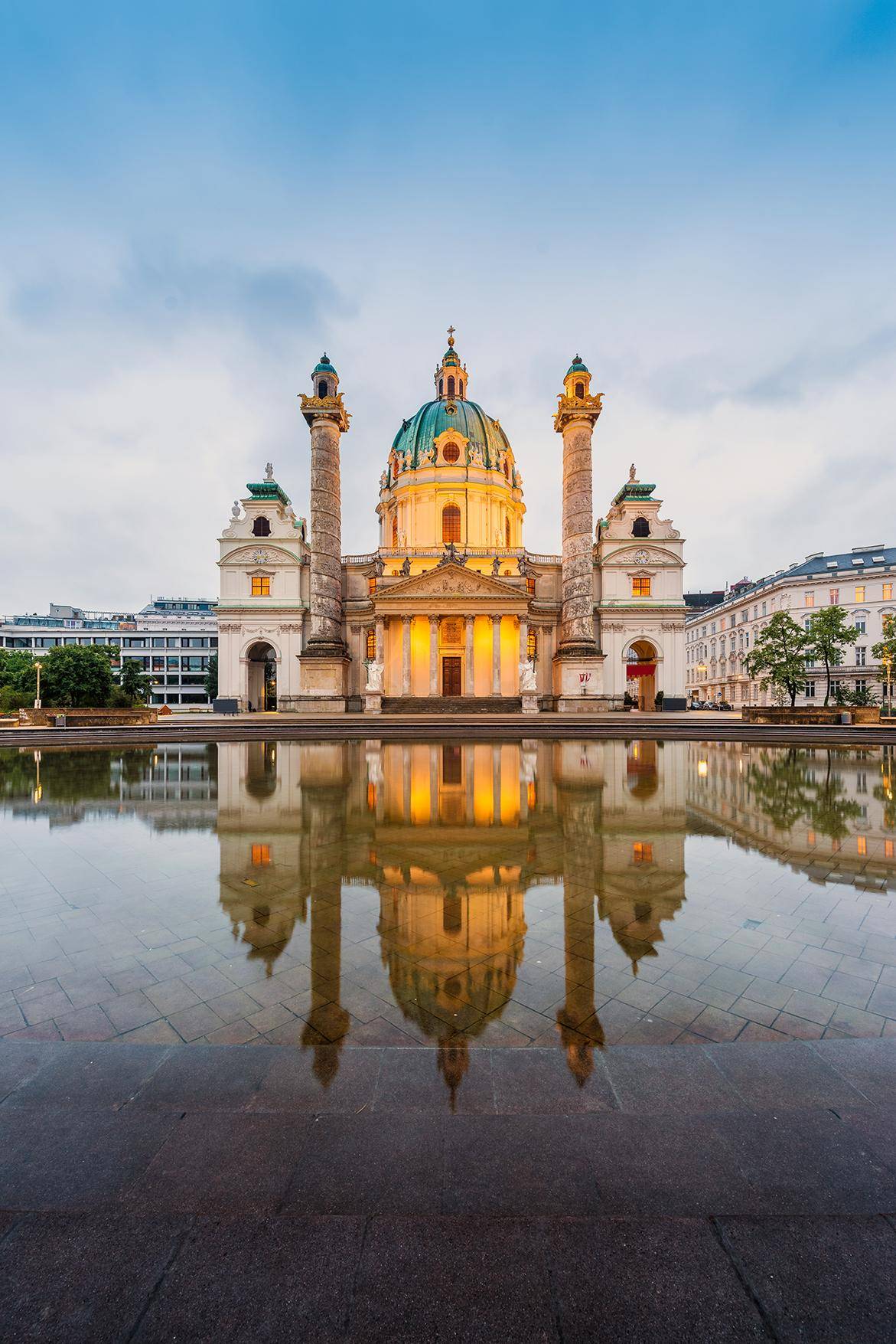 Abendliches Stadtbild der beleuchteten Karlskirche mit ihrem berühmten Brunnen in Wien. Der Himmel ist klar.