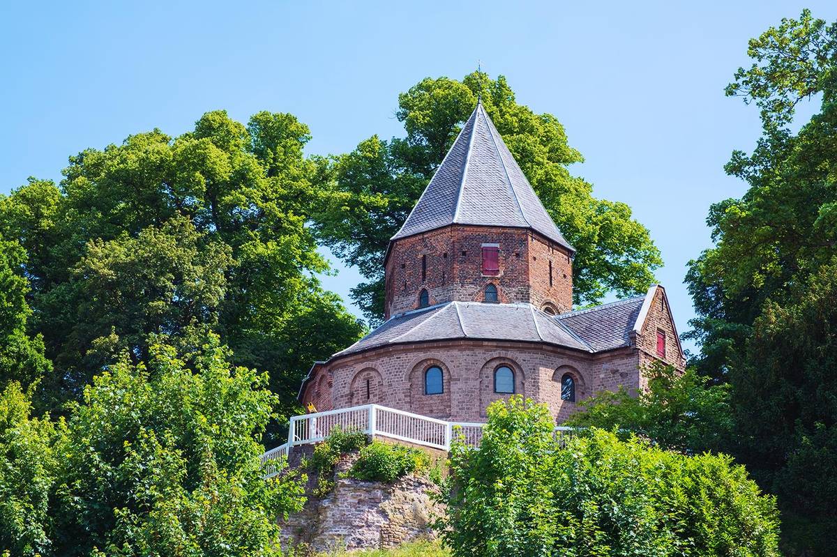 Die St. Nicolaas Kapelle blickt zwischen Bäumen und Büschen in einem Park in Nijmegen hervor. Man sieht das historische Gebäude von der Seite.