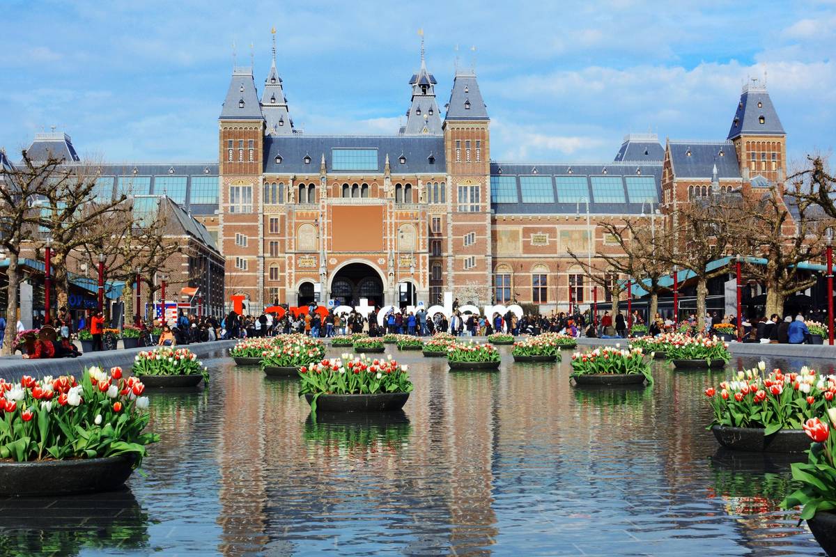 Das Historische Reichmuseum in Amsterdam. Vor dem Gebäude ist ein Brunnen mit Pflanzen, die sich im Wasser spiegeln. Es herrscht reges Treiben vor dem beliebten Museum