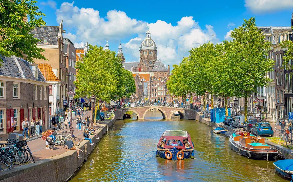 Herrliche Grachten in der Altstadt von Amsterdam. Man sieht eines der bekannten Tourenboote die regelmäßig Besuche zu Grachtenfahrten locken. Am Ende der Gracht erstrahlt ein historischer Kirchturm. Es tummeln sich einige Menschen an der Gracht.