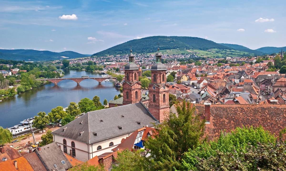 Historisches Stadtbild von Miltenberg. Man sieht die alte Brücke, den Kirchturm und die Dächer der Altstadt. Das Stadtbild ist von Backsteinbauten geprägt.