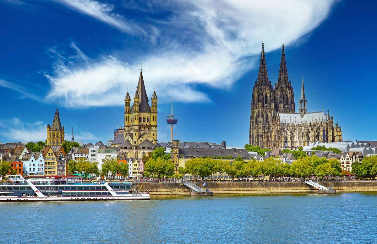 Ausblick auf die schöne Skyline von Köln. Zu sehen sind die Martinskirche und der bekannte Kölner Dom bei strahlend blauem Himmel. Am Rheinufer liegen Ausflugsboote an den Anlegestellen an.