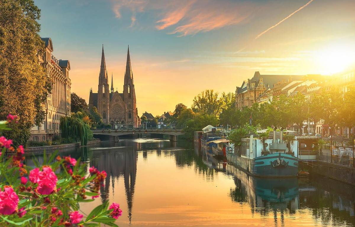 Aussicht auf Notre Dame in Straßburg. Die Kathedrale spiegelt sich durhc das goldene Licht der warmen Abendsonne im Waller. Am Ufer liegen Hausboote an.