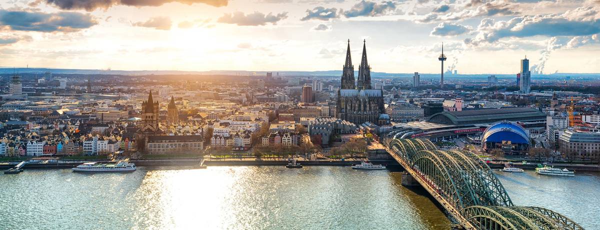 Schöner Ausblick auf die Kölner Rheinskyline. Man sieht den Kölner Dom, die Rheinuferpromenade, die Eisenbahnbrücke und das den Musical Dome bei abendlichem Sonnenlicht.